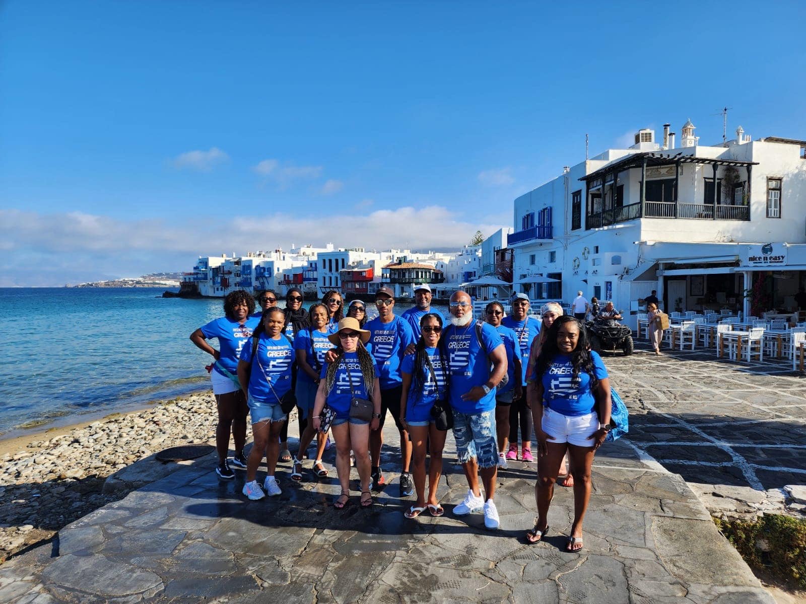 A Majestic Dream Vacations group in matching Greece shirts, gathered on the waterfront at Little Venice in Mykonos.