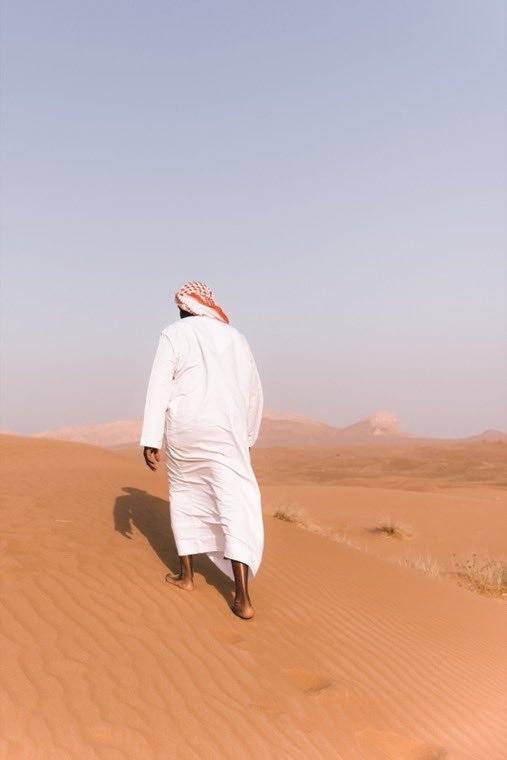 A figure in white robes walking out across desert dunes at dawn.