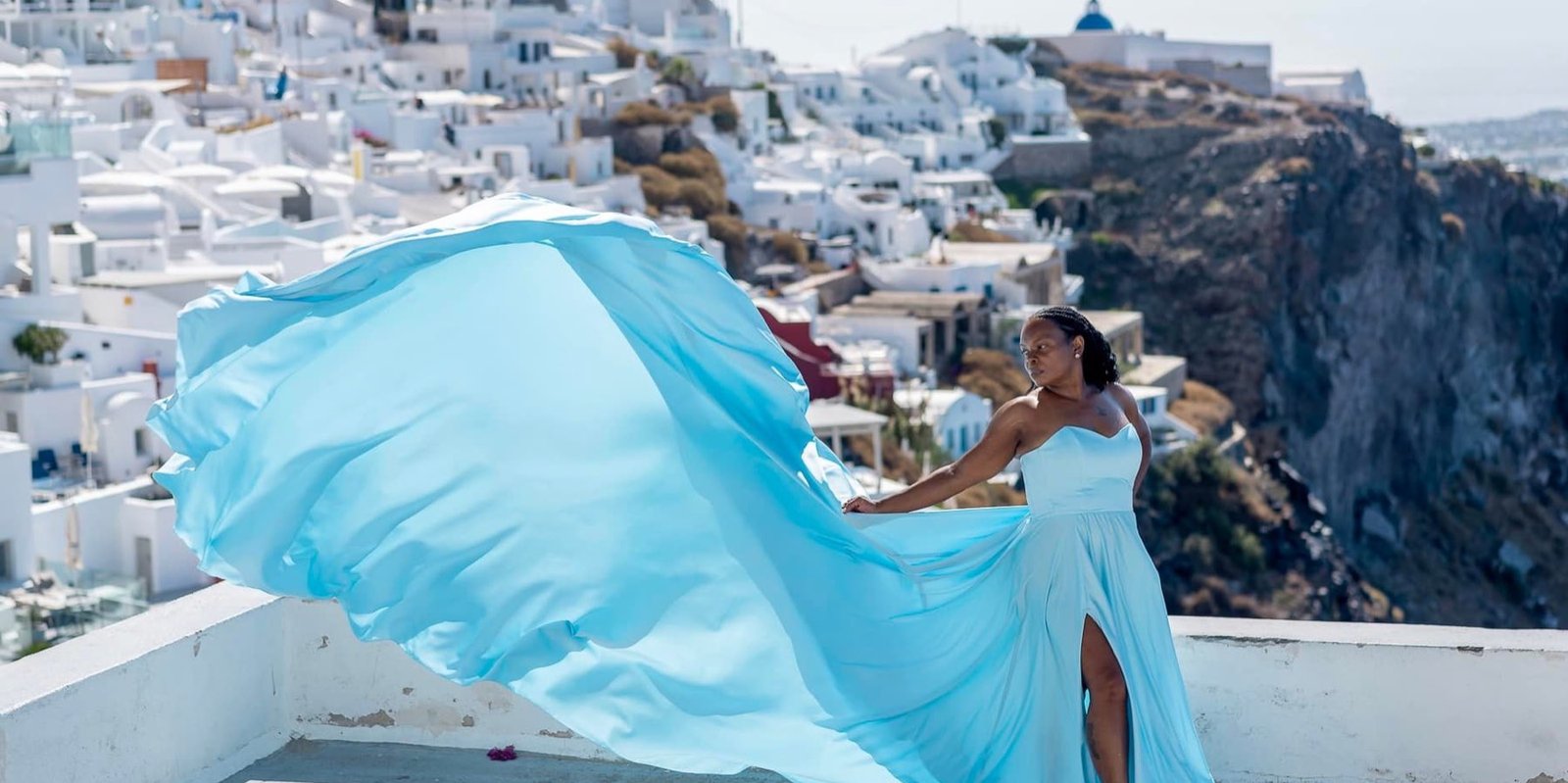 A traveler on a Santorini terrace in a pale blue gown, the long train caught by the wind against the white village behind.