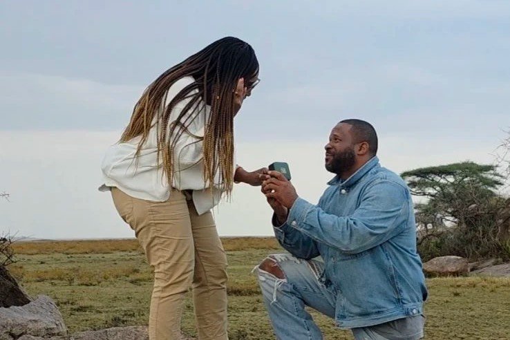 A marriage proposal on a rock outcrop in the savannah, one traveler kneeling with a ring box as the other looks down at it.