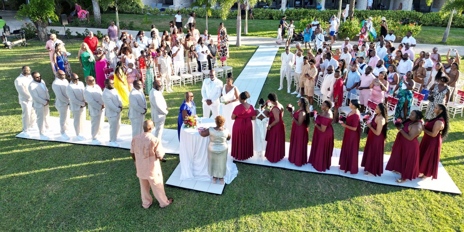 A destination wedding ceremony on a resort lawn, the couple at the front with the wedding party and around a hundred guests gathered on either side of the aisle.